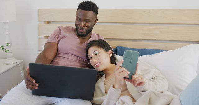 Happy diverse couple using laptop and lying in bedroom. Spending quality time at home concept.