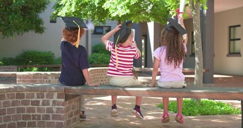 Joyful kindergarten graduates on bench wearing mortarboards holding diplomas in courtyard