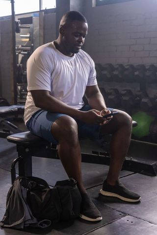 African american man sitting on workout bench checking smartphone after strength training