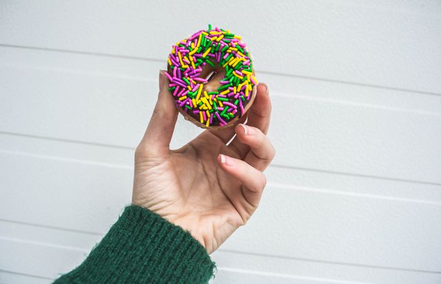 Hand holding colorful sprinkle doughnut close-up. Perfect for illustrating articles on desserts, sweets, or showcasing bakery products. Ideal for food blogs, social media promotion, and marketing materials for cafes or bakeries.