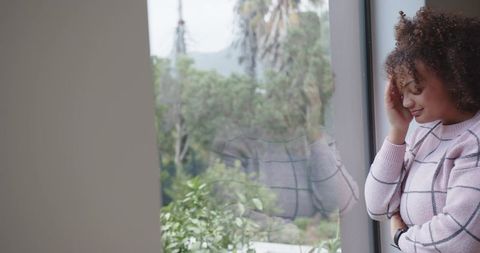 Contemplative woman leaning by window with soft reflection, pastel sweater, greenery