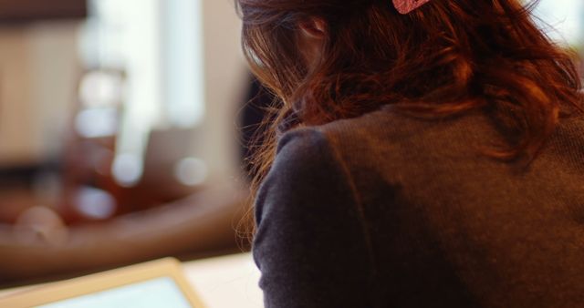 Young woman with long brown hair tied with a clip is leaning forward while reading on an e-book reader. The indoor environment offers a relaxed and modern setting with chairs and natural light. Ideal for use in technology-related themes, educational contexts, or lifestyle and leisure promotions.