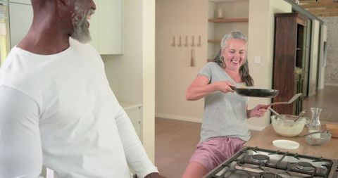 Senior couple laughing while cooking breakfast in cozy modern kitchen sharing pancake flip