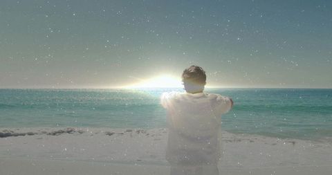 Man standing facing ocean at sunrise on sandy beach with calm teal seascape horizon