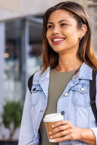 Smiling african woman holding coffee cup outside urban cafe with backpack and denim jacket, daylight