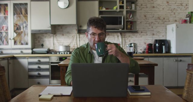 Middle-aged man working from home in a casual kitchen setting, drinking coffee while using his laptop. Ideal for themes related to remote work, home offices, telecommuting, productivity shots, work-life balance, and everyday life routines.