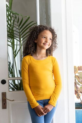 Smiling girl wearing mustard long-sleeve top standing by glass door with houseplants