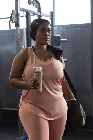 African american woman wearing pink activewear carrying gym duffel holding water bottle in gym