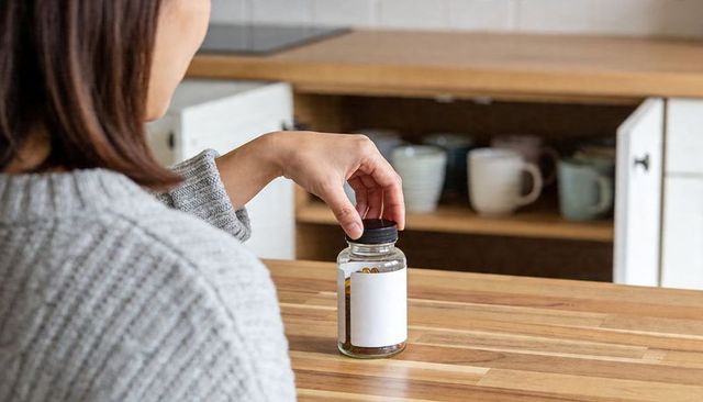 Asian woman reaching for amber supplement jar on wooden kitchen counter, capsule bottle