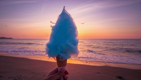 Hand holding blue cotton candy on waffle cone at sunset beach pastel sky seagulls