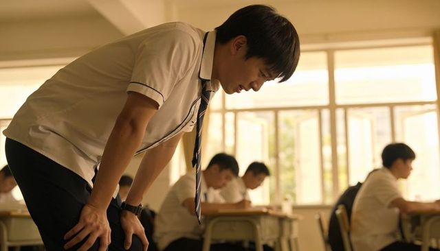 Teen student leaning over desk writing exam in sunlit classroom during focused study