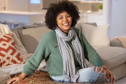 Smiling woman sitting on sofa in green sweater and gray scarf, cozy home interior