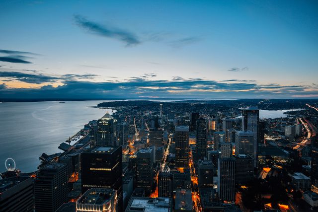Aerial view of a bustling city at twilight, showcasing buildings illuminated by glowing lights. Ideal for urban planning presentations, travel advertisements, and marketing materials emphasizing modern metropolitan life. The dramatic evening sky complements the cityscape, creating a vibrant and dynamic visual perfect for backgrounds, websites, and postcards.