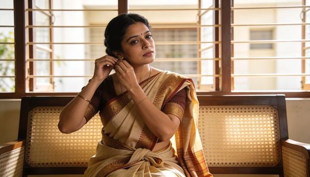 Indian woman adjusting earrings wearing cream gold saree sitting on cane bench by window