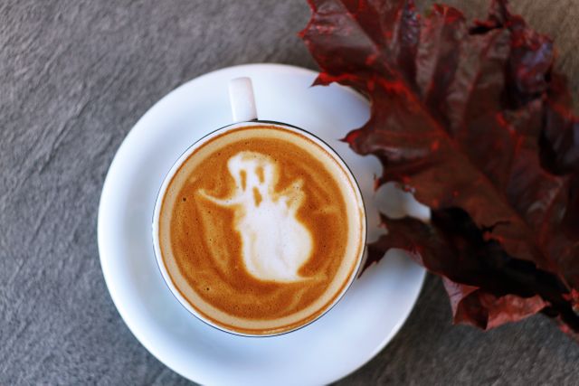 Latte art features a ghost design, creating a spooky atmosphere perfect for Halloween or autumn-themed content. The autumn leaf beside the cup adds a seasonal touch. Ideal for promoting seasonal coffees, cafes, holiday events, and festive blog posts.