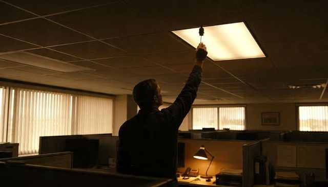 Maintenance worker adjusting fluorescent ceiling panel in dim open-plan cubicle workspace
