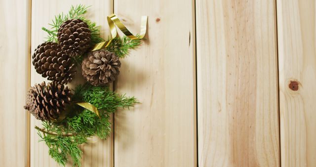 Pinecones and green foliage arranged on wooden planks with a gold ribbon. Ideal for holiday decorations, nature-themed projects, rustic designs, and festive event planning. This can be used for creating greeting cards, seasonal marketing materials, or as a simple natural background for websites or print media.
