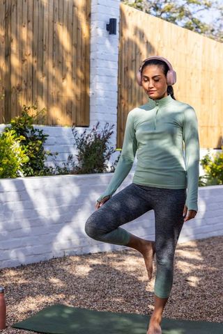 Indian woman practicing tree pose on green yoga mat in backyard wearing pink headphones