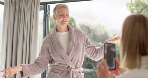 Mature woman posing in plush bathrobe while younger woman recording on smartphone