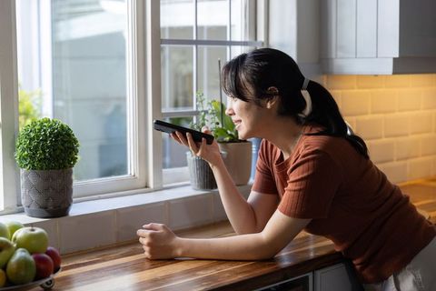 Asian woman leaning on kitchen counter speaking into smartphone by window with plants