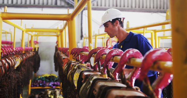 Worker sorting metal hooks and industrial equipment in manufacturing plant. Man is wearing blue coveralls and a safety helmet, focusing on organizing tools. Useful for industrial workplace presentations, safety training material, and illustrating manual labor in factories.