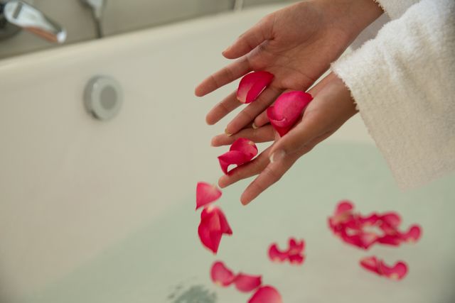 Mid section of biracial woman spending time at home self isolating and social distancing in quarantine lockdown during coronavirus covid 19 epidemic, throwing rose petals in bathtub relaxing.