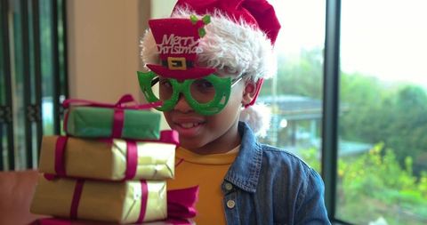 African american boy wearing santa hat and green glasses holding stacked christmas gifts