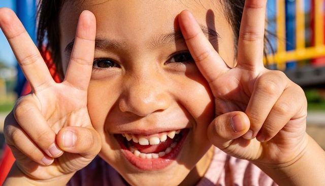 Sunlit asian girl smiling and making v-signs at colorful playground close-up portrait
