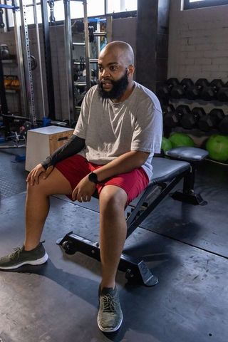 African man sitting on bench pausing during gym workout wearing gray tee and red shorts