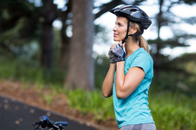 Female cyclist adjusting helmet while standing on a road in a forested area. Ideal for use in advertisements promoting cycling safety, outdoor activities, fitness, and healthy living. Suitable for blogs, websites, and magazines focused on sports, exercise, and active lifestyles.