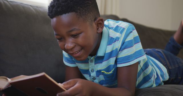African american boy reading a book while lying on the couch at home. staying at home in self isolation