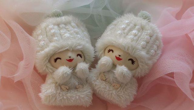 Matching plush twin dolls wearing knit hats on pastel tulle, cozy cuddly nursery decor