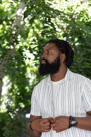 Adult african man adjusting striped shirt in sunlit garden with dreadlocks and beard
