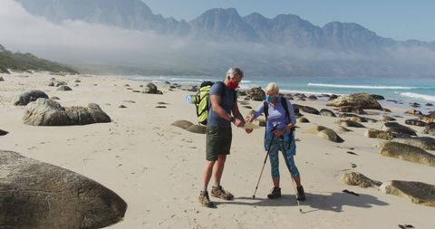 Senior Couple Hiking on Beach with Masks and Backpacks