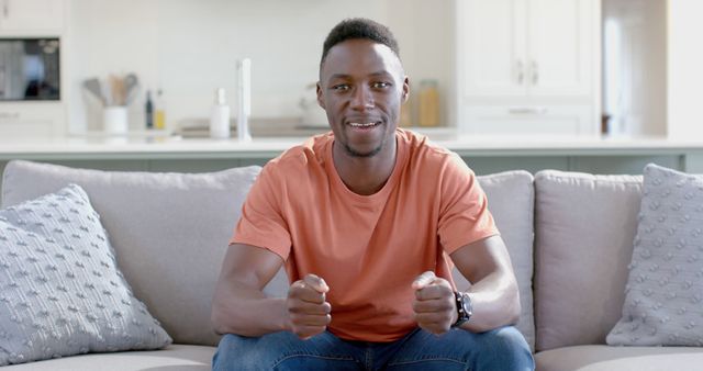 Young man enthusiastically watching sports on television, sitting on a comfortable couch in a modern living room. Suitable for promoting sports events, television services, or casual living lifestyle.