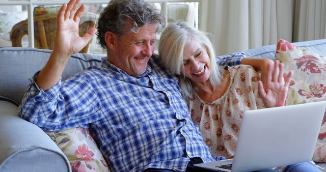 Senior couple sitting on a couch, smiling and waving to a laptop, indicating they are video calling loved ones. The cozy home and casual attire suggest comfort and warmth, making this perfect for use in advertisements for communication technology, family connections, or retirement lifestyle content.