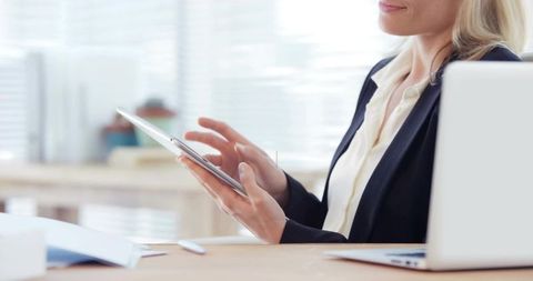 Professional woman tapping tablet at desk with laptop and paperwork, modern daylight office