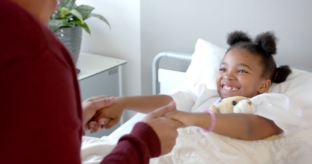 Happy african american girl patient and her mother holding hands in bed in hospital room. Medicine, healthcare and hospital, unaltered.