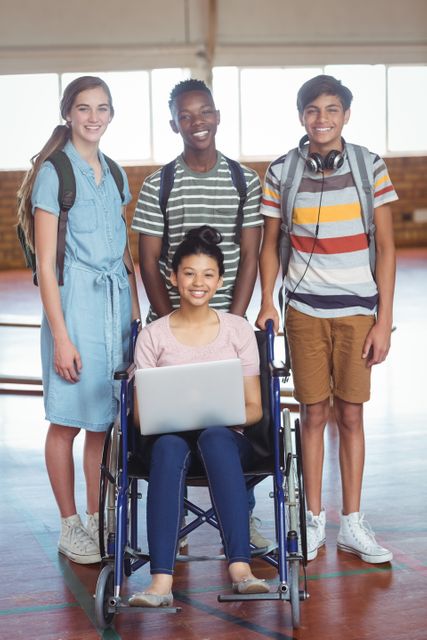 Diverse group of students standing in a school gym, smiling and looking at the camera. One student is in a wheelchair holding a laptop, while others have backpacks. Ideal for use in educational materials, inclusivity campaigns, and youth-oriented content.