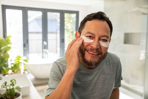 Mature asian man applying under-eye gel patches at bathroom vanity smiling, self-care routine