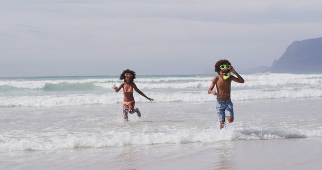 Two children running happily on a sandy beach with ocean waves in the background. Both are dressed in swimwear, reflecting the joy of a summer vacation. The image also features mountains on the horizon. This can be used in travel brochures, family vacation advertisements, or summer activity promotions.