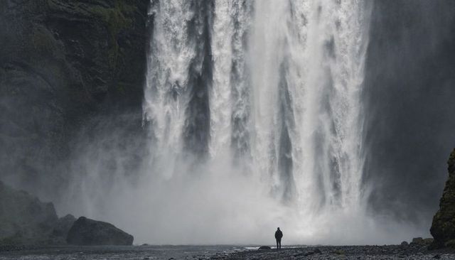 Solitary figure facing massive cascading waterfall on misty pebble riverbank