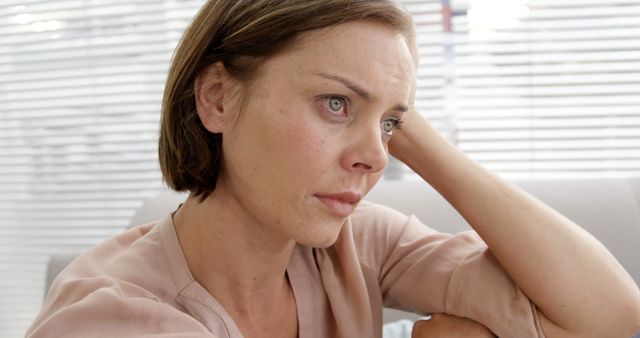 A woman with short hair, dressed in casual clothes, sits indoors by a window with blinds, looking away and appearing deep in thought. This visual is perfect for themes surrounding contemplation, emotional moments, mental health awareness, loneliness, or reflection. It can be used in blog posts, articles, or advertisements relating to personal experiences, psychology, or wellness.