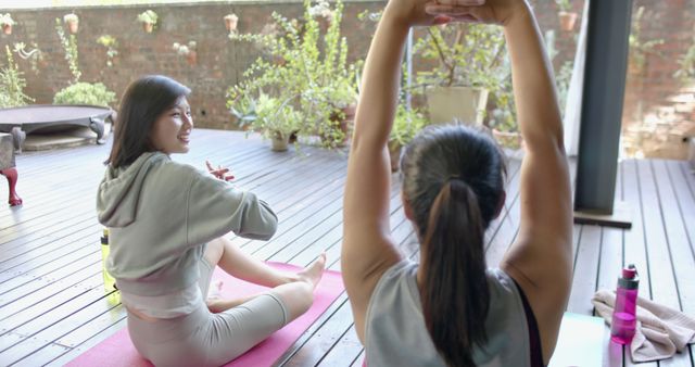 Two women practicing yoga together on a wooden deck surrounded by plants. Great visual for promoting wellness, outdoor activities, fitness sessions, healthy lifestyle content, and exercise routines.