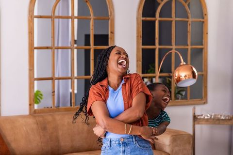 African mother and teenage daughter laughing and hugging on cozy living room sofa