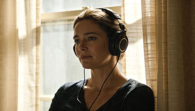 Contemplative woman wearing headphones leaning by sunlit window with sheer curtains