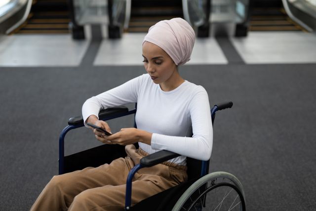 Young biracial woman using smartphone in wheelchair. She has light brown skin, wearing pink headscarf and casual clothes, unaltered