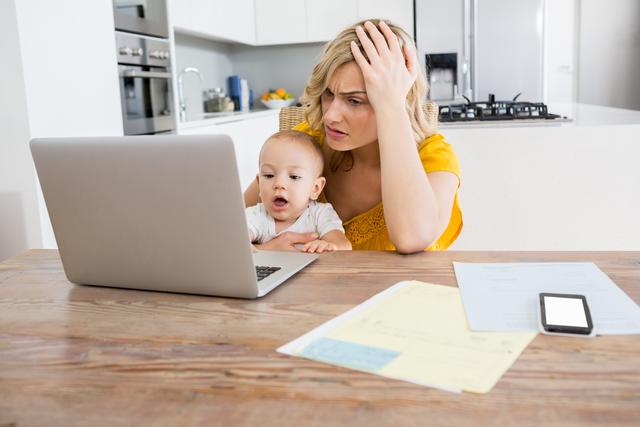 Mother is working on a laptop while holding her baby in a modern kitchen. She appears stressed, possibly due to balancing work and parenting responsibilities. This image is ideal for articles or advertisements related to work-life balance, parenting challenges, remote work, and family life.