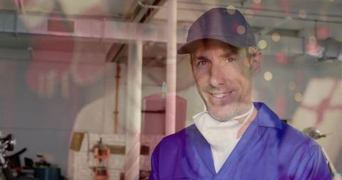 Smiling mechanic wearing cap and respirator working in motorcycle garage with bokeh lights