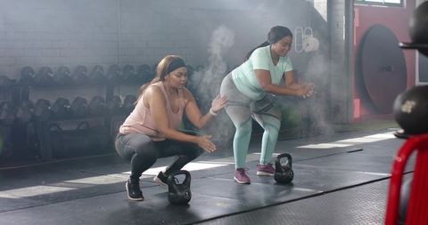 Diverse women performing synchronized kettlebell squats with chalk cloud in fitness studio